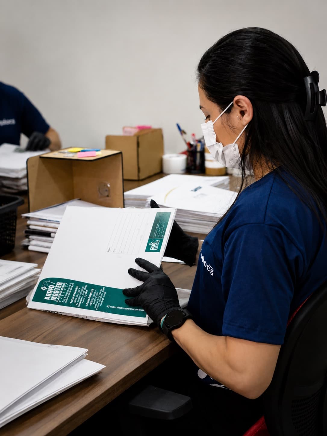 Arquivista da SOS Docs, de máscara e luvas, conferindo folha a folha um maço de documentos na bancada de preparação, com outros lotes empilhados na mesa e a equipe trabalhando ao fundo