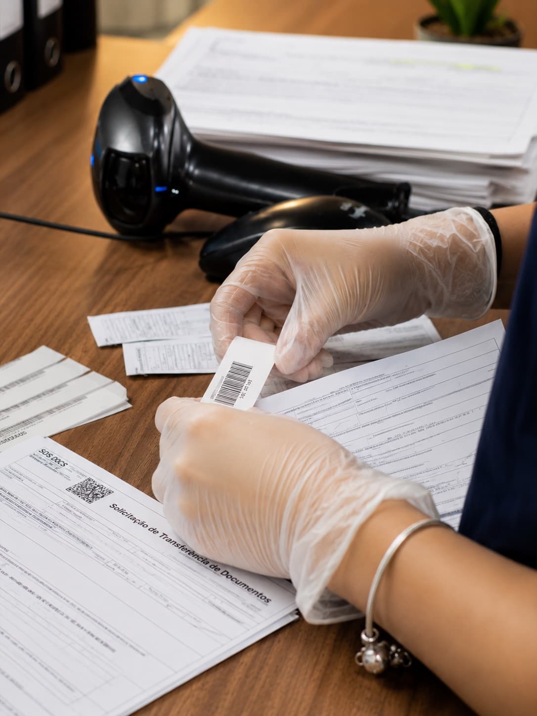 Mãos com luvas aplicando etiqueta de código de barras sobre a solicitação de transferência de documentos, ao lado do leitor óptico